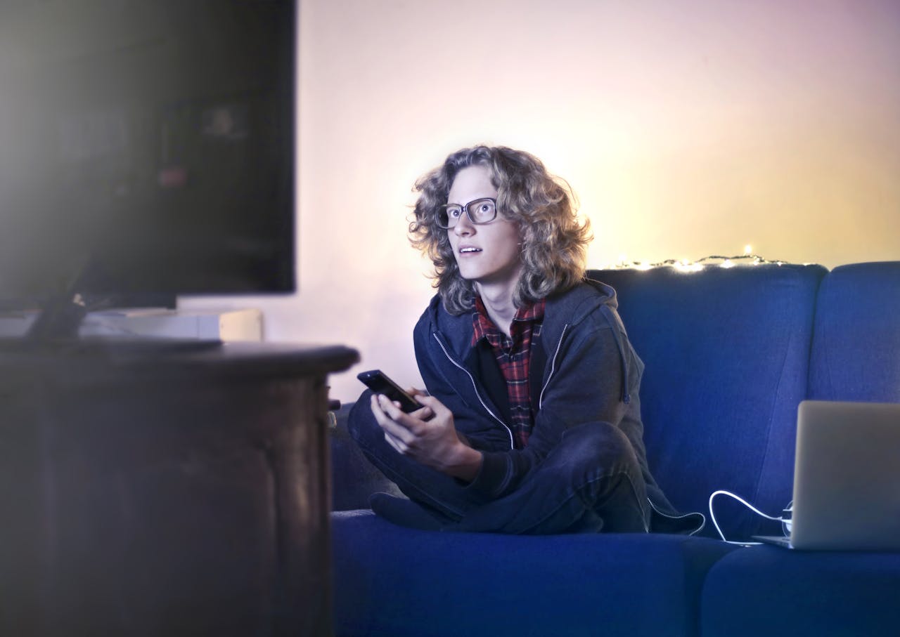 about-us Concentrated male with long hair sitting on comfortable sofa at home and messaging on social media via cellphone while watching movie on TV with opened mouth