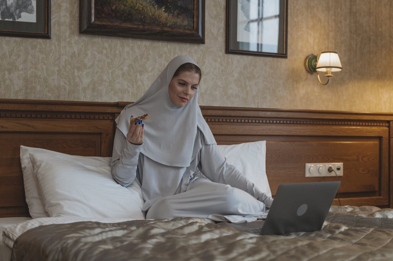 services-03 A Muslim woman in a hijab enjoys pizza while watching a laptop in a cozy bedroom setting.